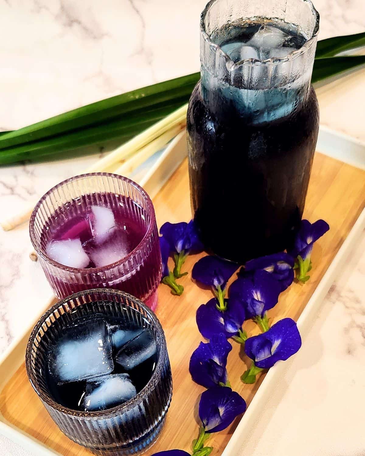 Two glasses of butterfly pea flower tea with one glass jar and some flowers on a wooden tray.
