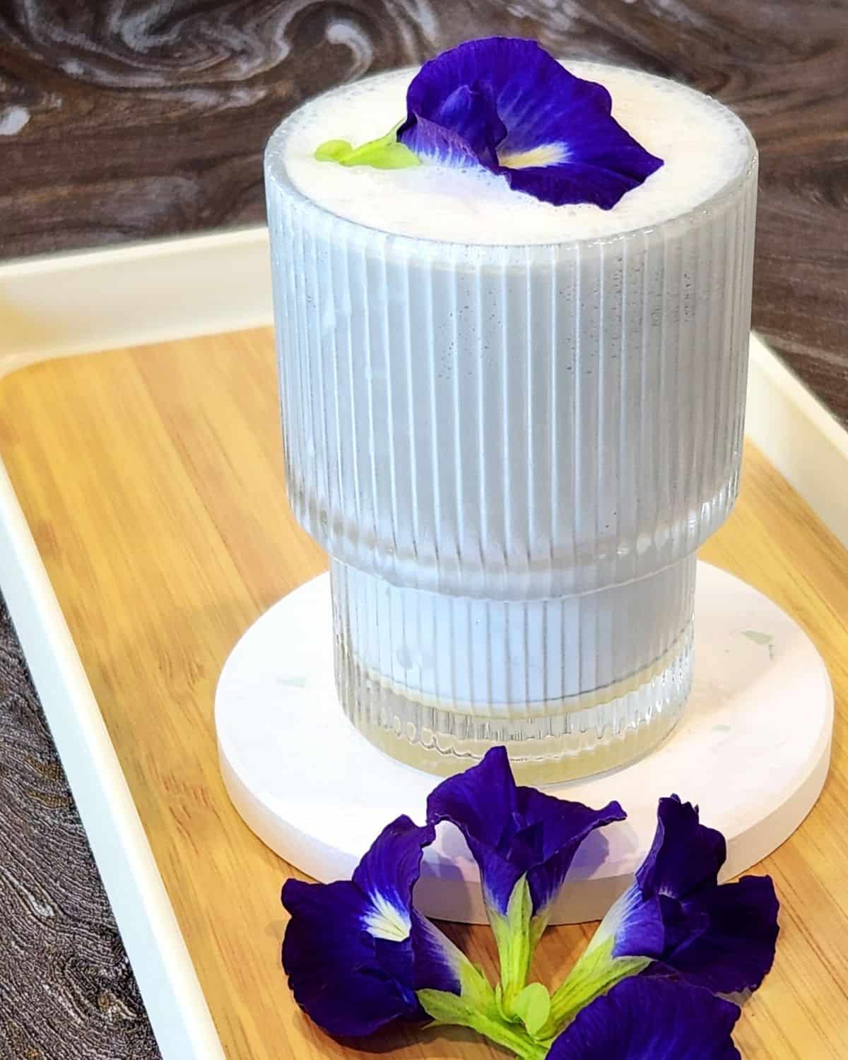 A glass of butterfly pea flower latte on a wooden tray with some fresh flowers on.