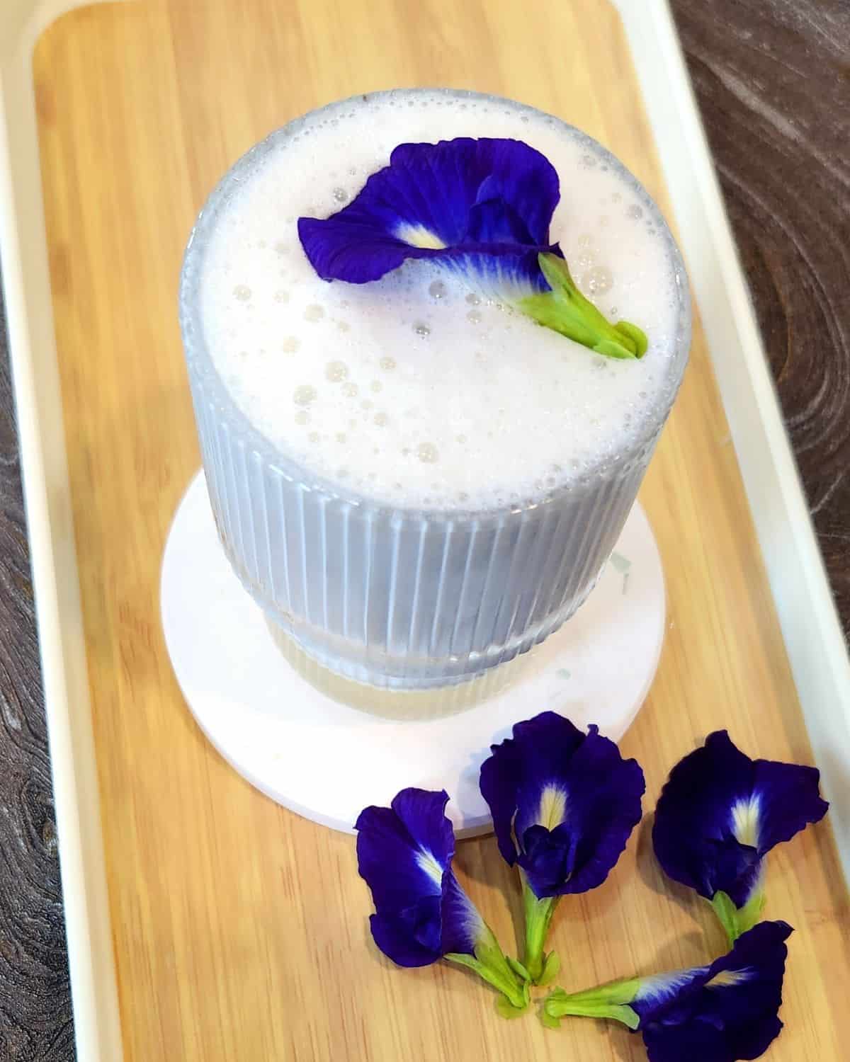 A glass of butterfly pea flower latte with fresh butterfly flowers on a wooden tray.