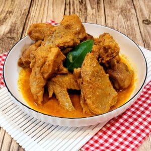 A picture of chicken rendang in a white plate on red white cloth.
