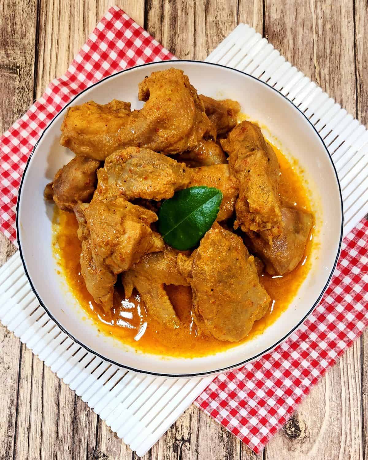 A photo of chicken rendang in a white plate with red white cloth on a wooden table.