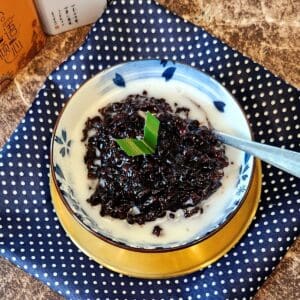 A bowl of pulut hitam porridge with coconut sauce on a golden plate and blue and white cloth.