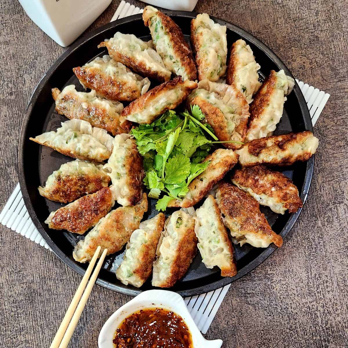 A photo of chicken potstickers lined on a round, black plate with coriander leaves in the center.