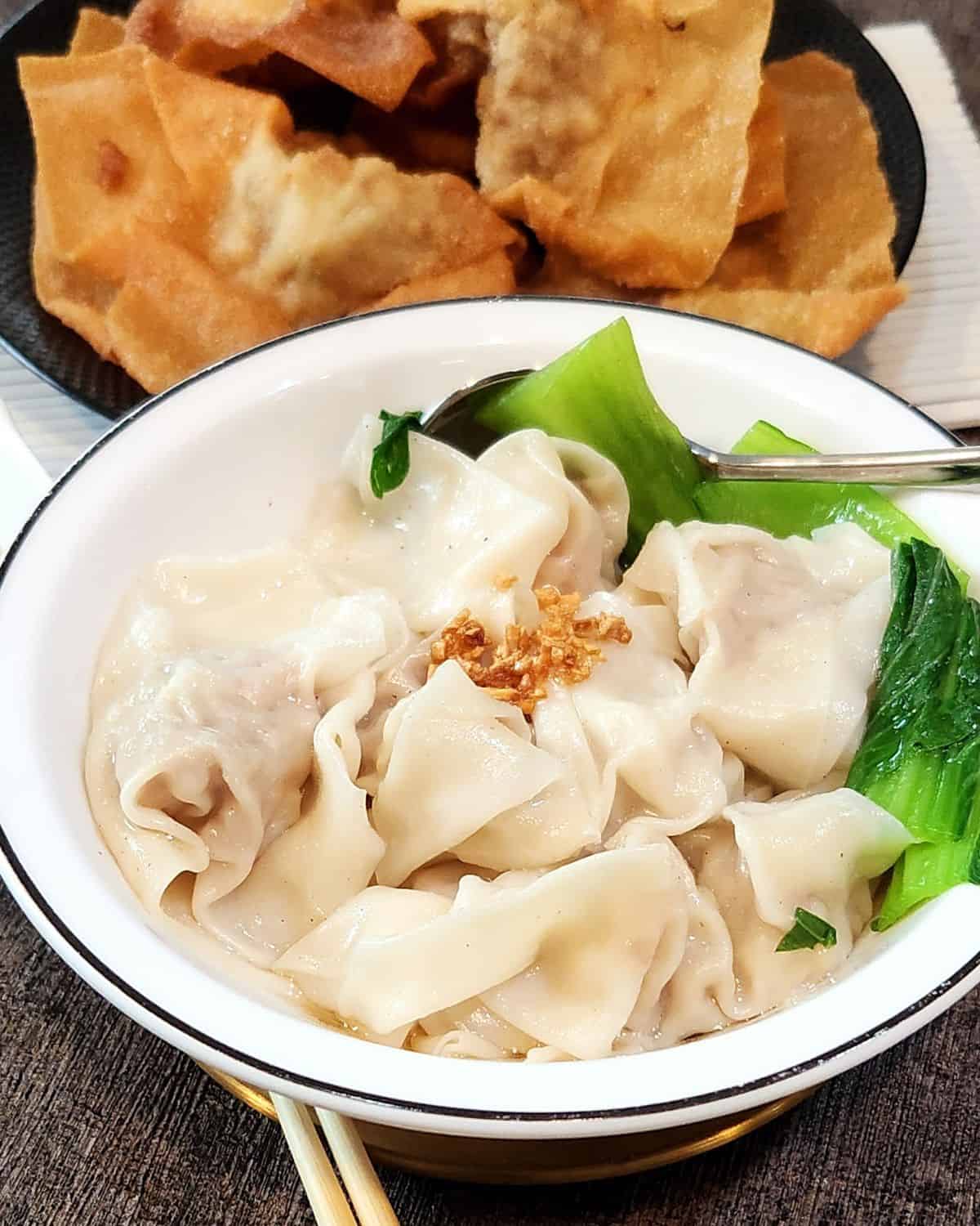 A bowl of boiled wontons soup with deep-fried wontons at the background.