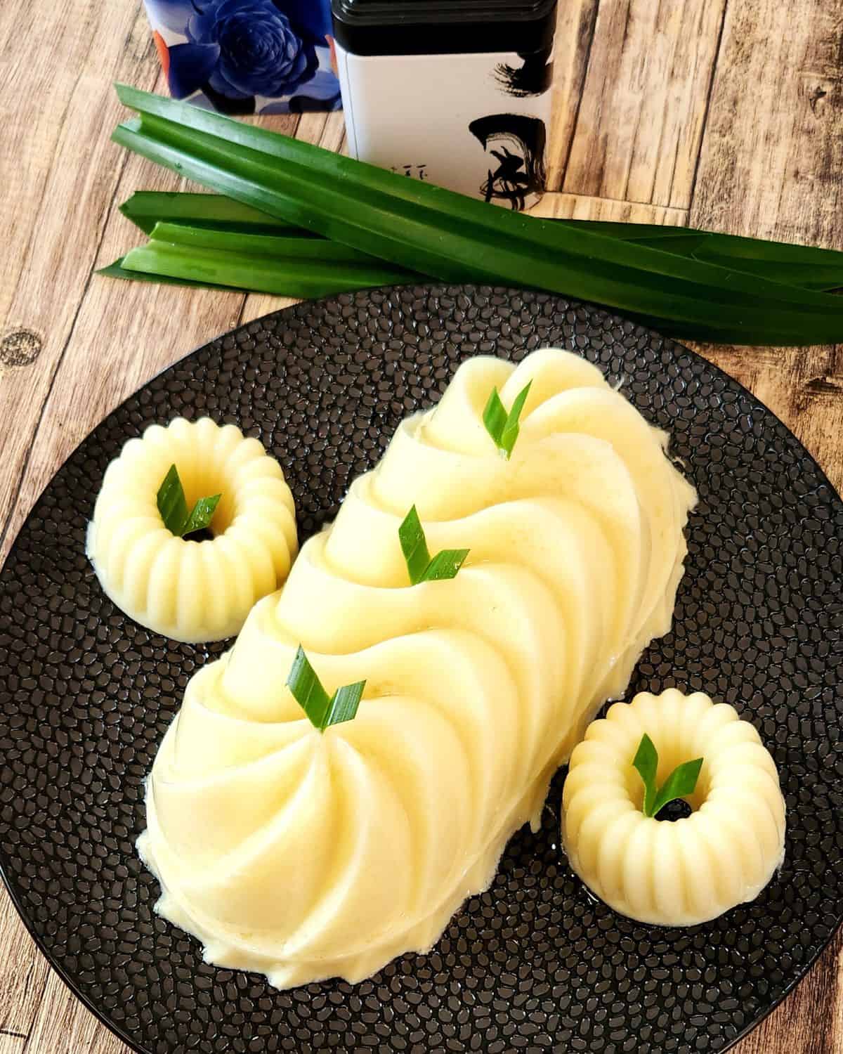 Three pieces of Gluten Free Corn pudding on a black plate with pandan leaves as decoration.