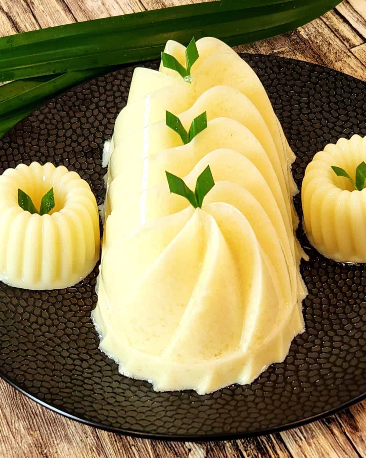 Three pieces of Gluten Free Corn pudding on a black plate with pandan leaves as decoration.