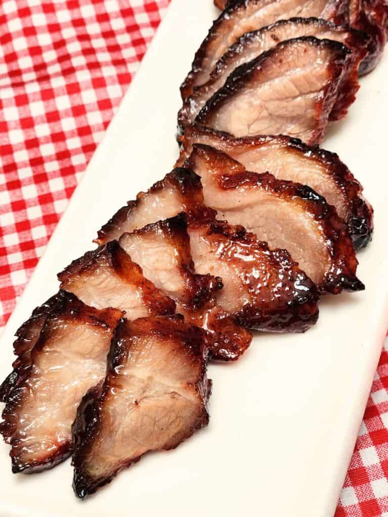 Char siu roast pork sliced on a long white plate is placed on red white table cloth.
