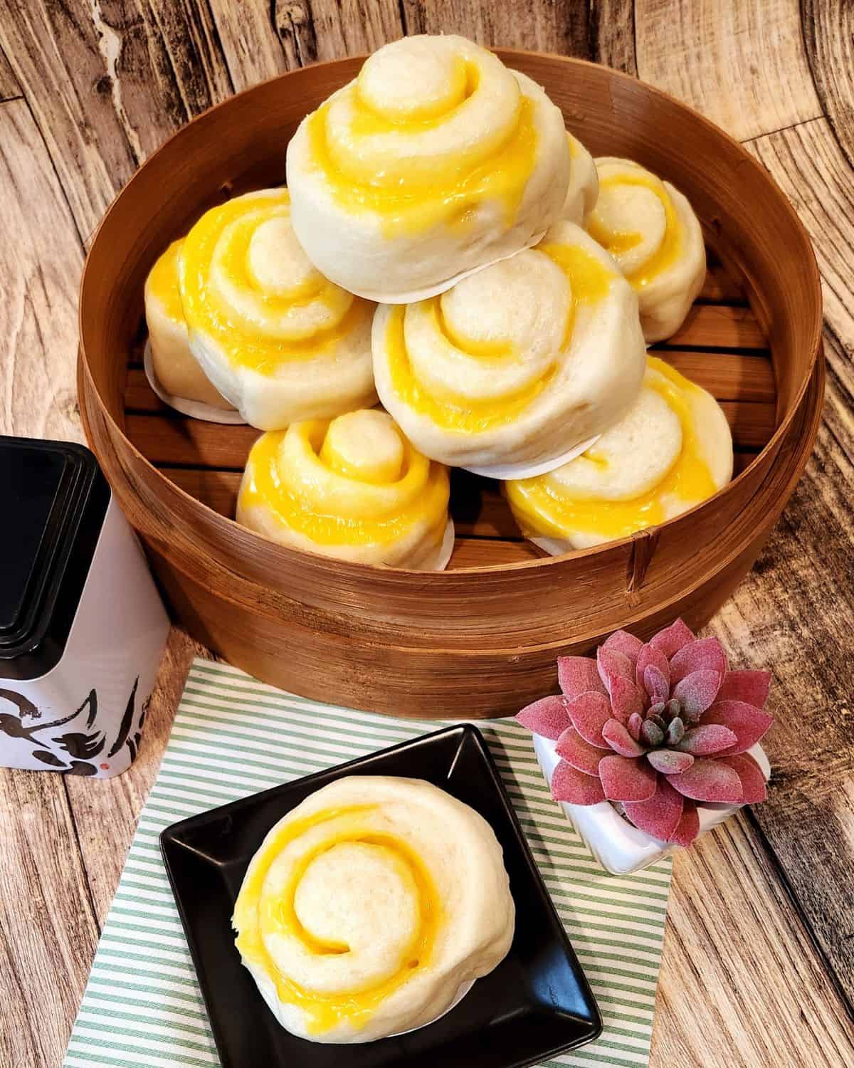 Chinese steamed bun with Cheese filling piled on a bamboo steamer and one bun on a small black plate next to it.