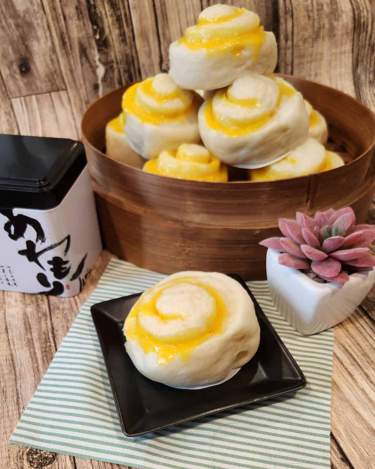 Chinese steamed bun with Cheese filling piled on a bamboo steamer and one bun on a small black plate next to it.