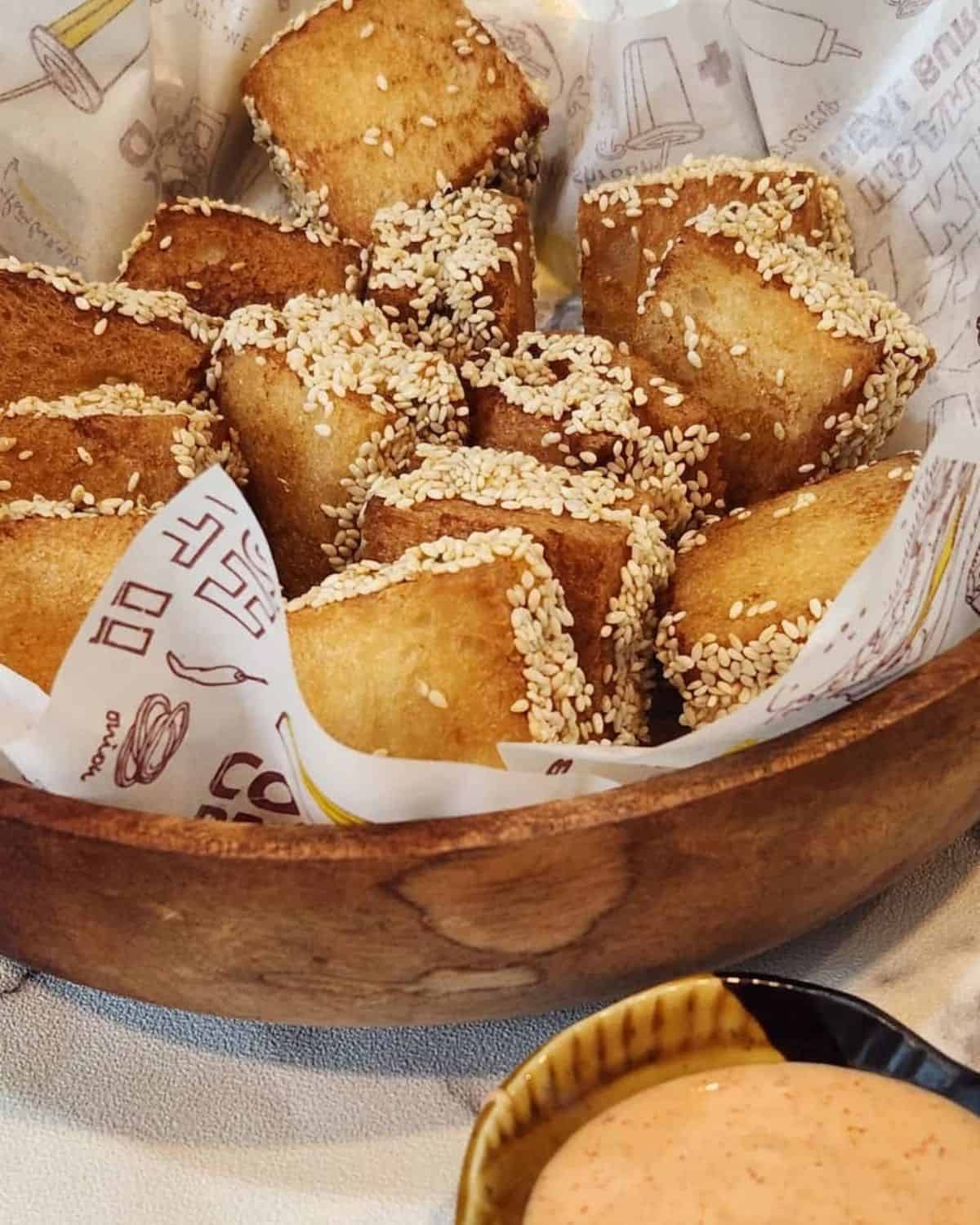 Several pieces of Menbosha - Korean fried shrimp toast in a wooden basket with menbosha sauce next to it.