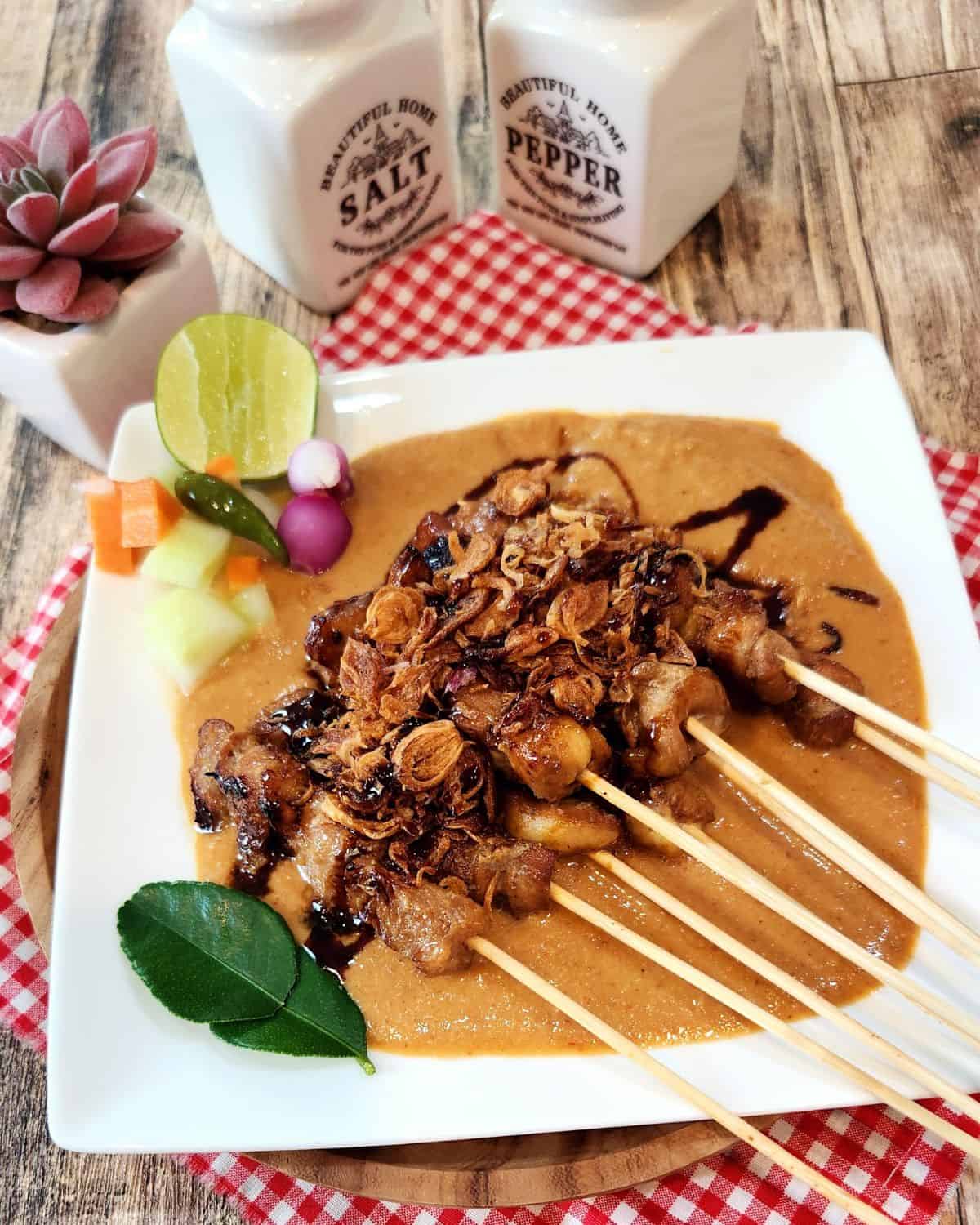A plate of chicken satay with peanut sauce, some pickled cucumbers, lime wedge, fried shallots.