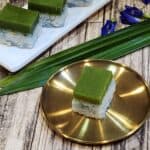 A golden plate of kuih seri muka with butterfly pea flowers and pandan leaves at the back.