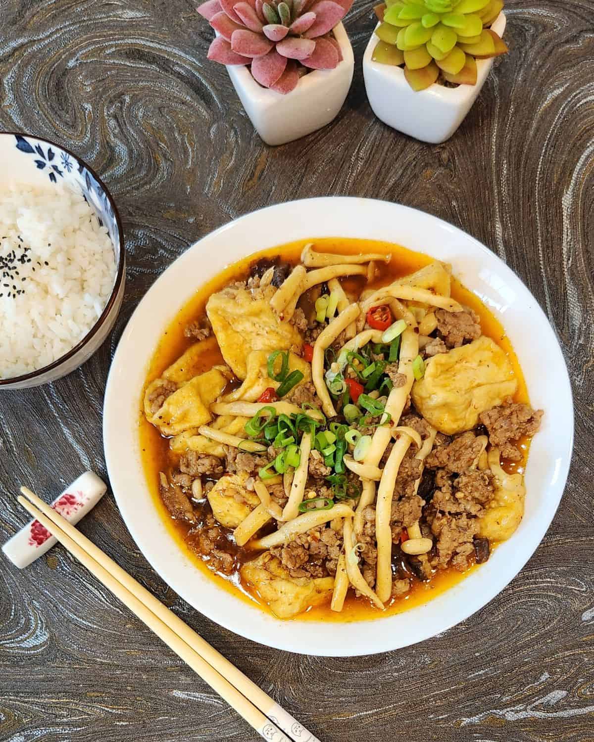 A plate of Szechuan tofu with mushrooms and ground pork with chopsticks and a bowl or rice.