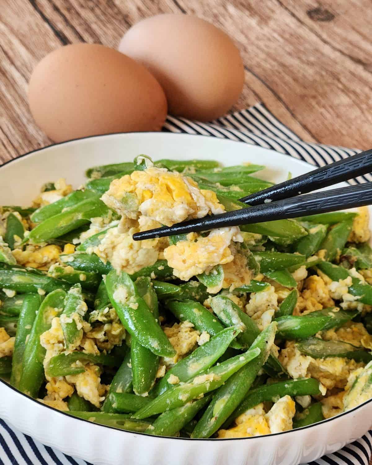 A pair of chopsticks holding green beans with eggs.