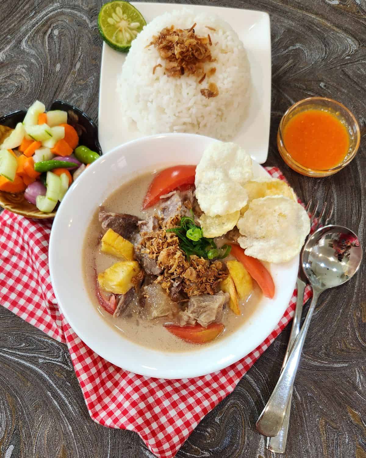 A bowl of Soto Betawi with white rice, chili sauce, and acar timun.