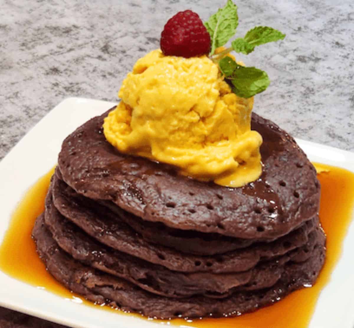 A stack of ube mochi pancakes drizzled with maple syrup and topped with mango ice cream.