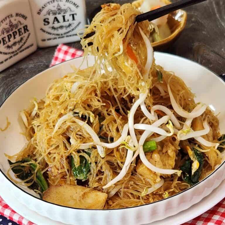A pair of chopsticks holding bihun goreng from a plate.