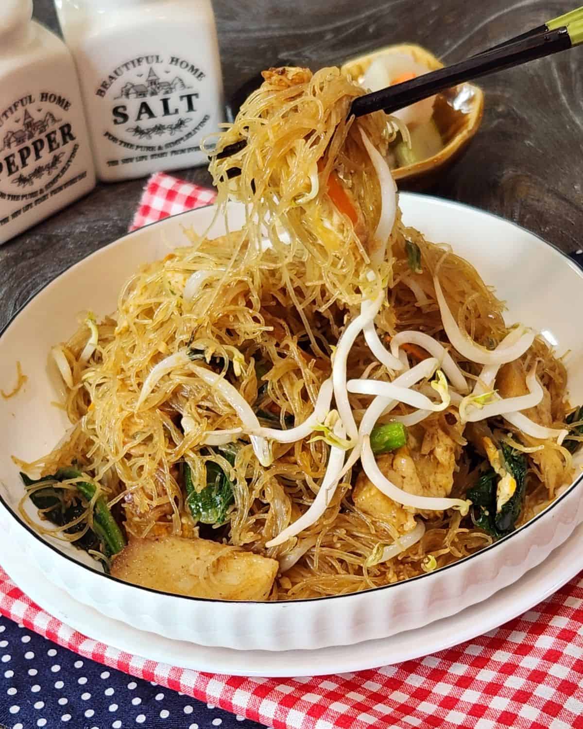 A pair of chopsticks holding bihun goreng from a plate.