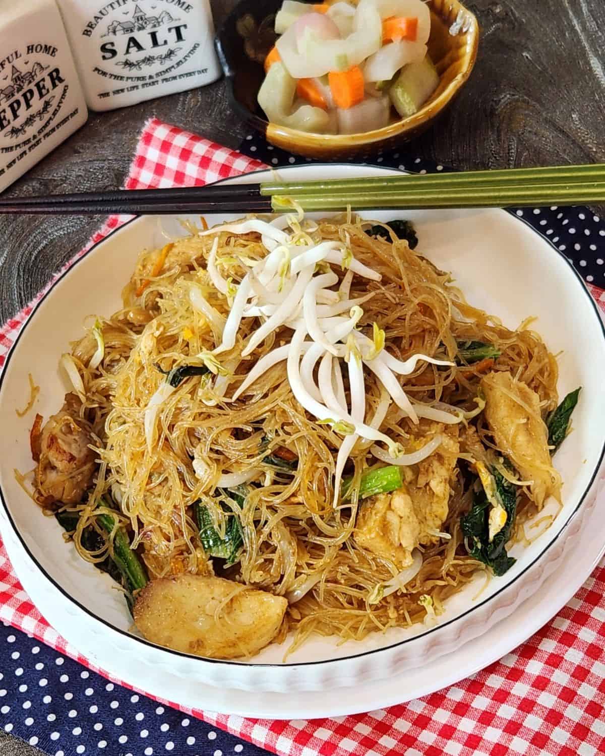 A plate of bihun goreng with chopsticks and pickled cucumber next to it.