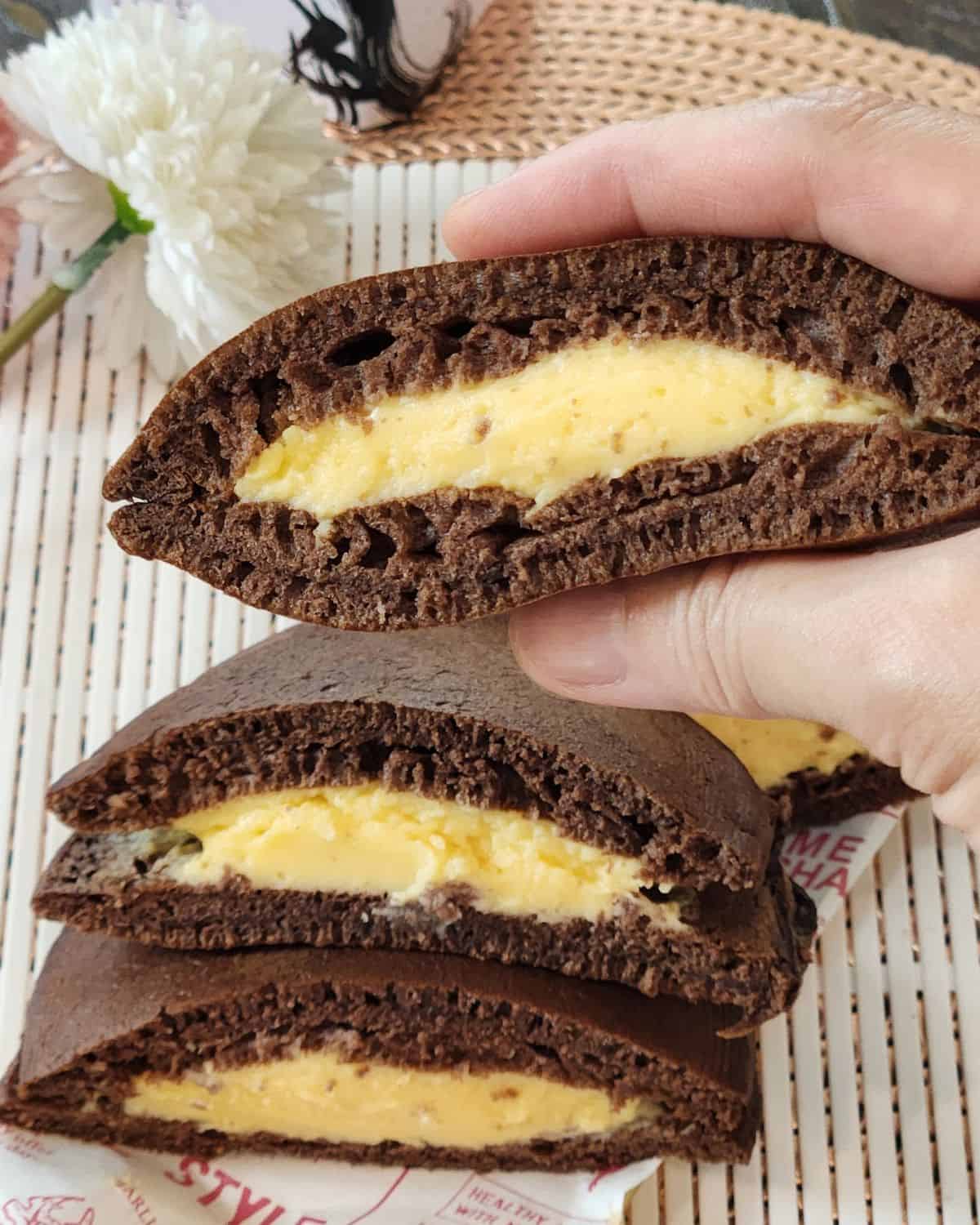 A hand holding the fluffy chocolate dorayaki with custard filling inside.