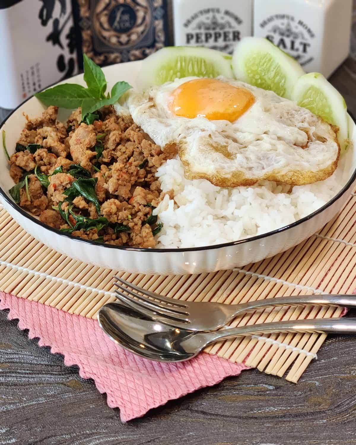Pad Kra Pao pork dish with crispy fried egg, holy basil, and fresh cucumber slices.