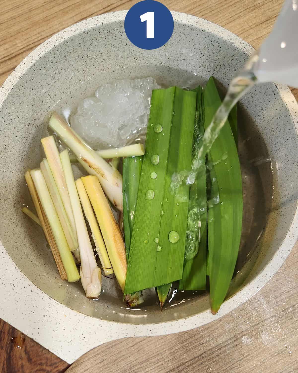 Lemongrass stalks, pandan leaves, and rock sugar in a pot with water pouring in.