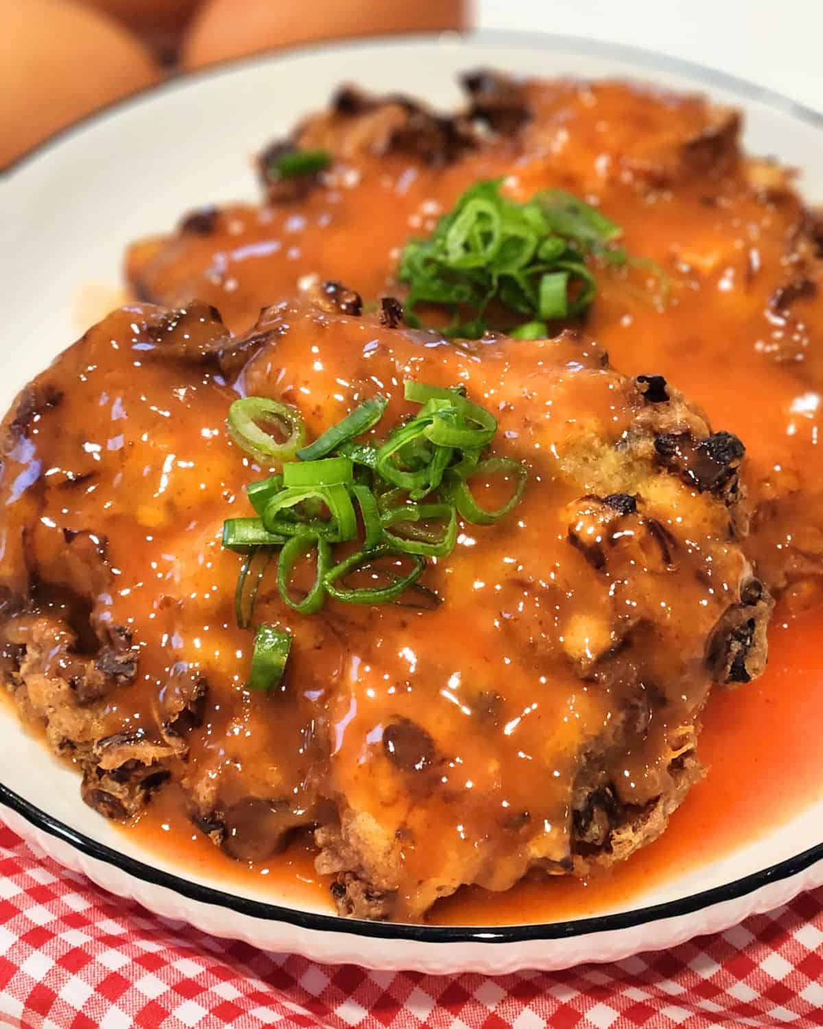 A plate of two pork and shrimp egg foo young patties with tomato sauce drizzled on top and scallions for garnish.