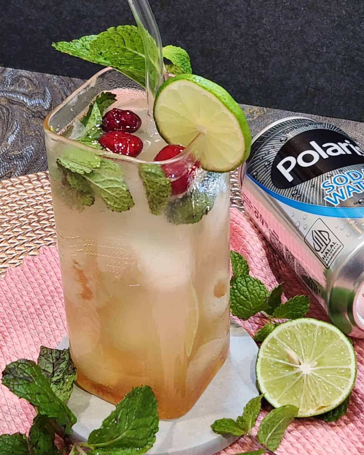 A glass of lychee soda with mint leaves, cranberries, lime, and ice cubes.