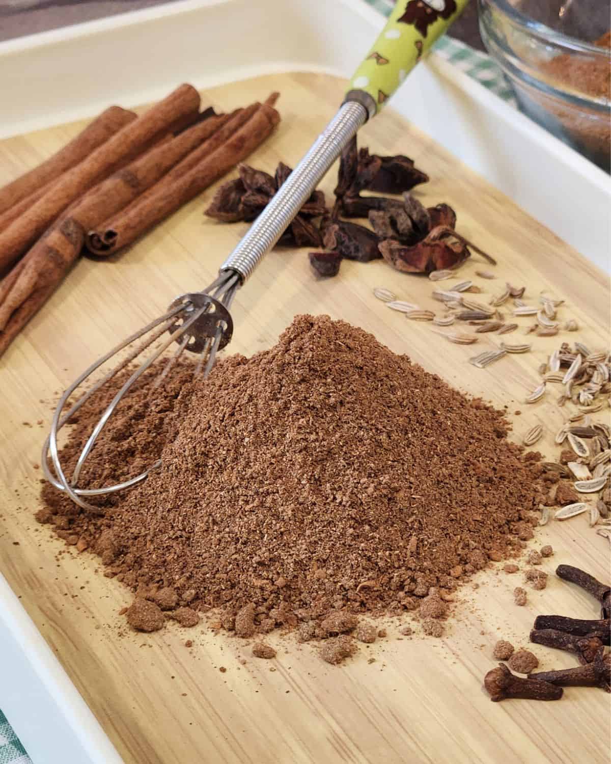 A stack of Chinese five spice powder with some spices behind and a small whisk.