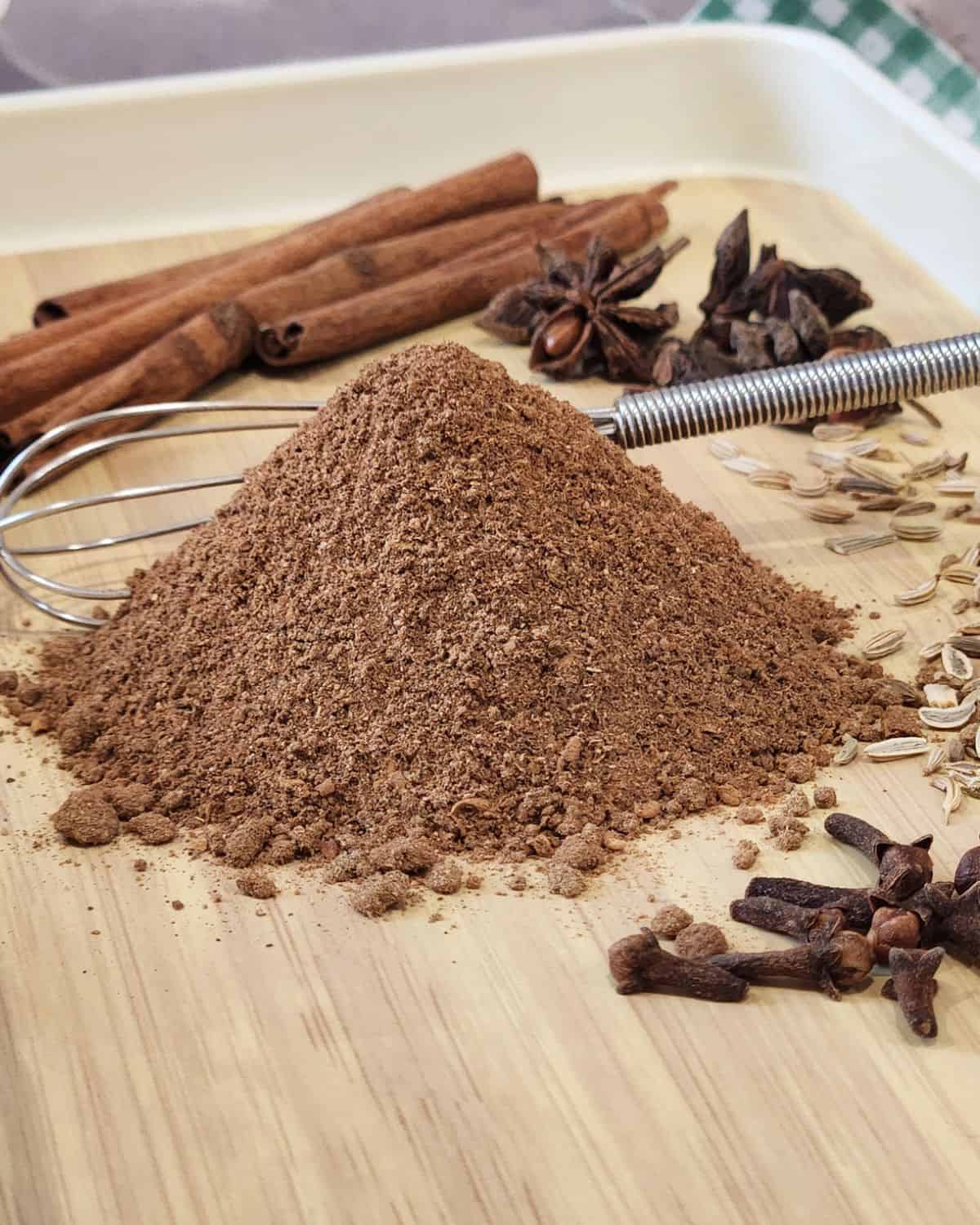 A stack of Chinese five spice powder with some spices behind and a small whisk.