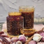 Two jars full of garlic chili oil with shallots, garlic, and herbs in front of them.