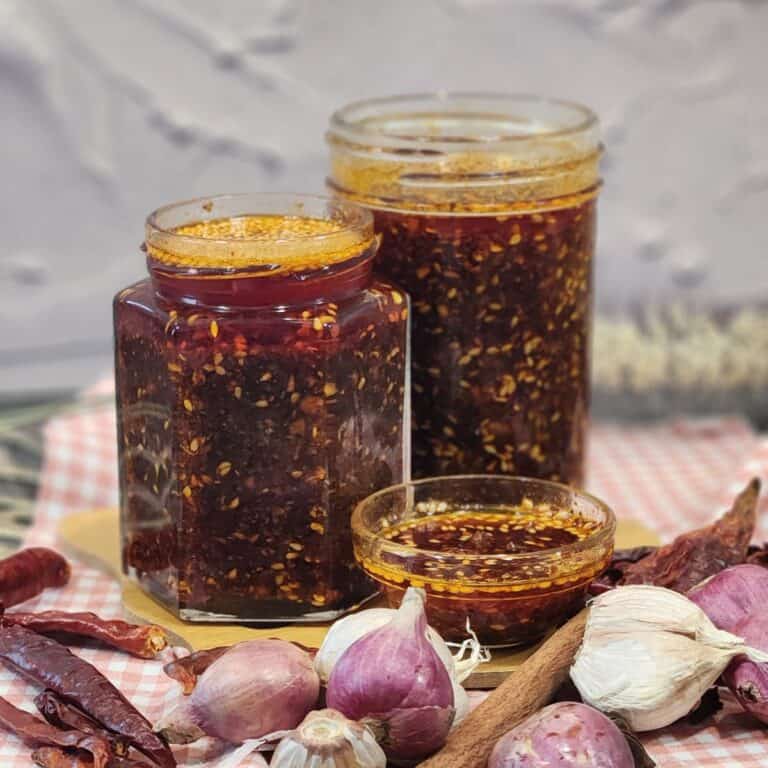 Two jars full of garlic chili oil with shallots, garlic, and herbs in front of them.