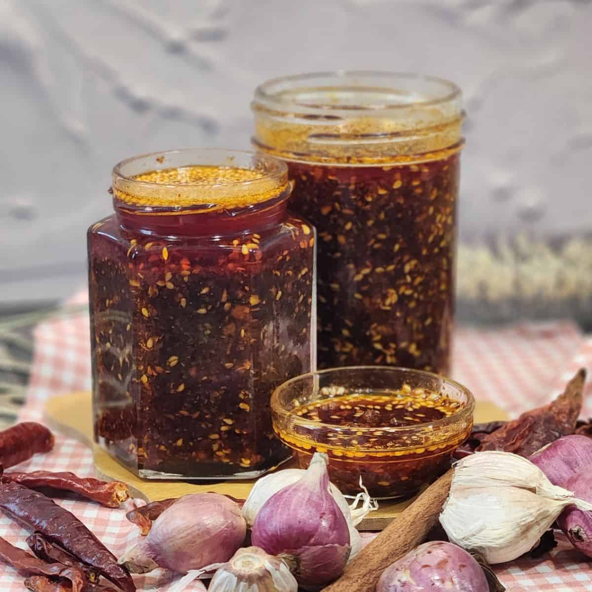 Two jars full of garlic chili oil with shallots, garlic, and herbs in front of them.