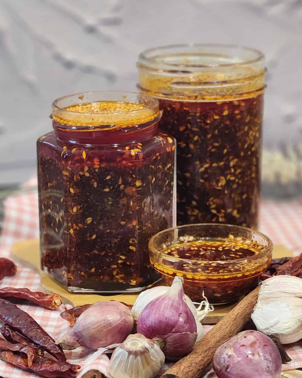 Two jars full of garlic chili oil with shallots, garlic, and herbs in front of them.