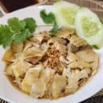 A plate of silky Hainanese Chicken drizzled with soy sauce, and garnished with cilantro and cucumber slices.