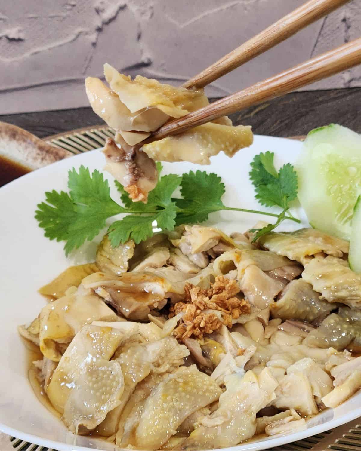 A pair of chopsticks holding a piece of Hainanese chicken drizzled with soy sauce mixture.