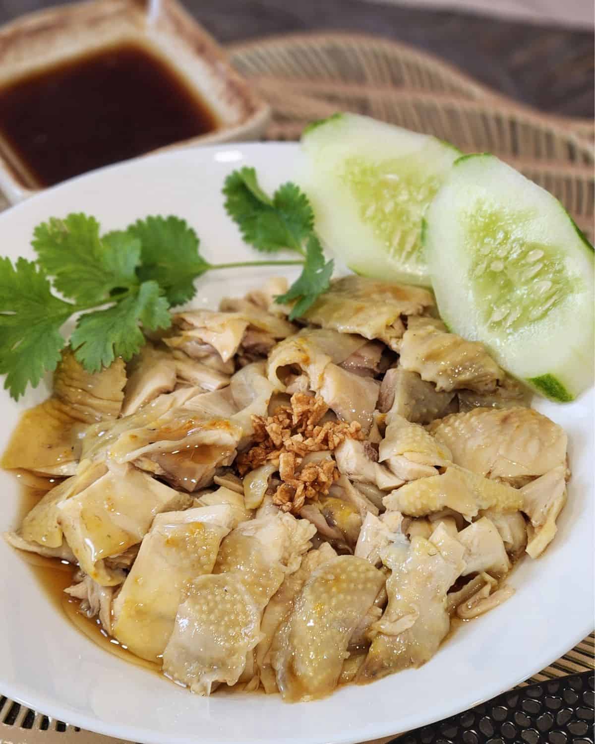 A plate of silky Hainanese Chicken drizzled with soy sauce, and garnished with cilantro and cucumber slices. 