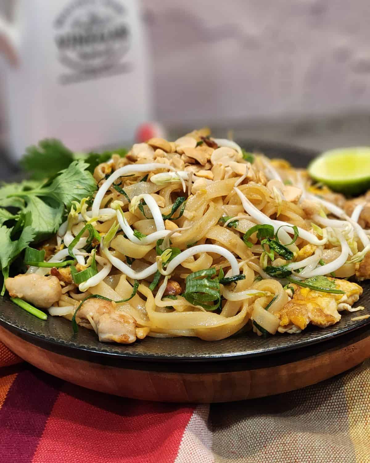 A black plate with Chicken pad thai in it, garnished with cilantro and lime wedges.