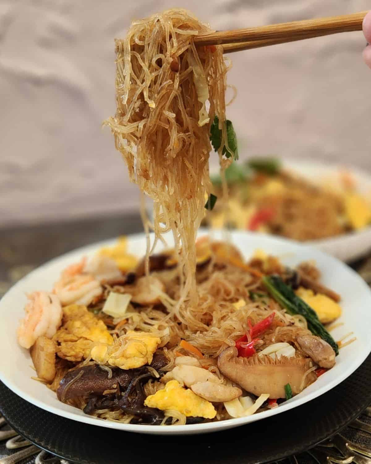 A pair of chopsticks holding stir fried glass noodles in the air.