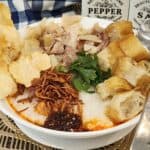 A bowl of chicken confee with toppings like shredded chicken, youtiao, chili oil, fried shallots, crackers, and celery.