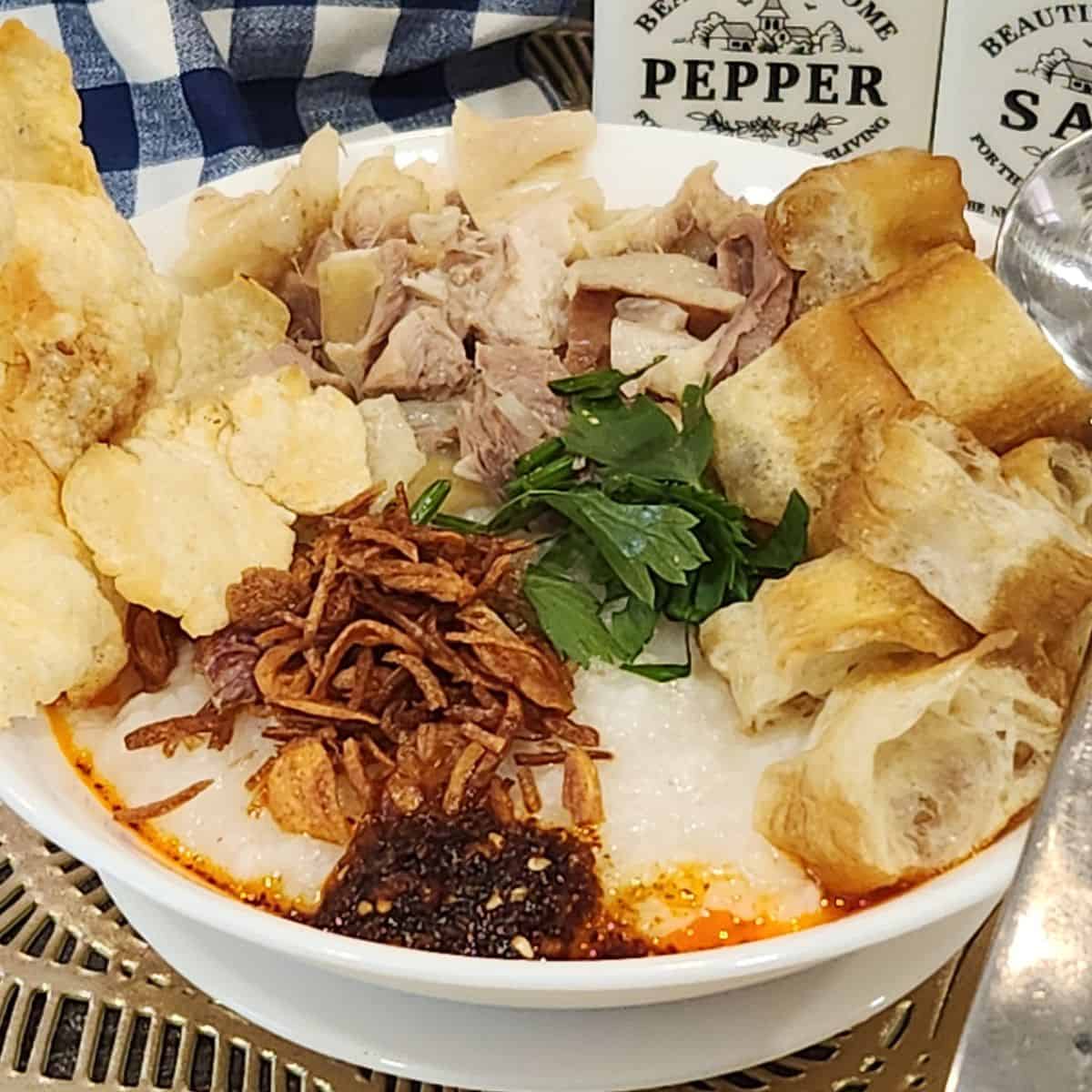 A bowl of chicken confee with toppings like shredded chicken, youtiao, chili oil, fried shallots, crackers, and celery.