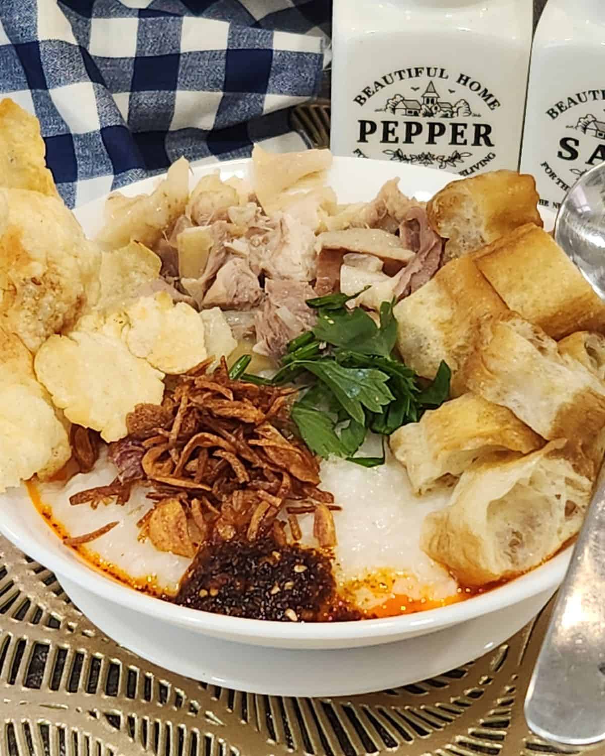 A bowl of chicken confee with toppings like shredded chicken, youtiao, chili oil, fried shallots, crackers, and celery.