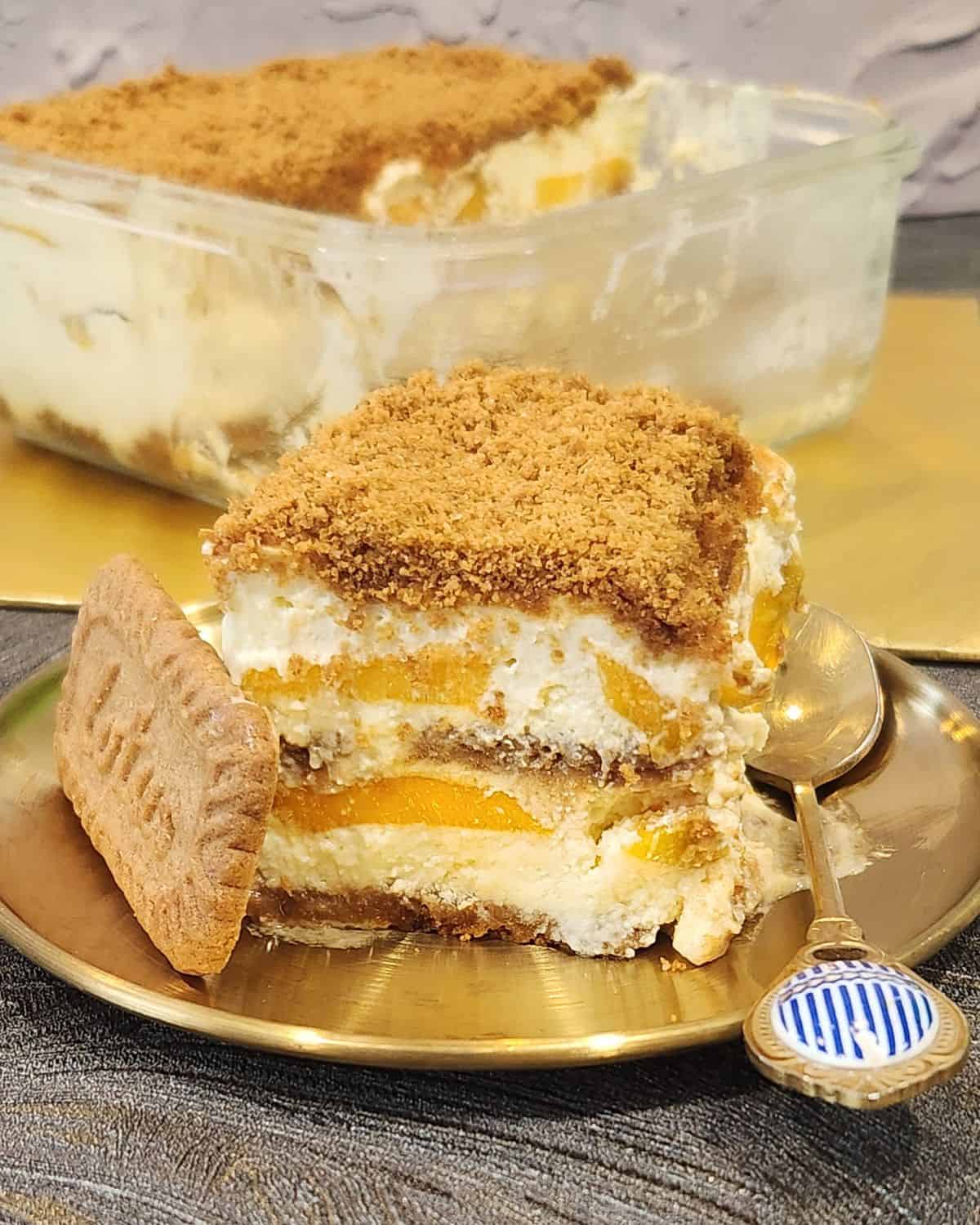 A square piece of mango float on a golden plate with a piece of lotus biscoff biscuit.